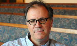 A middle-aged man with short brown hair and round black glasses is sitting indoors. He is wearing a blue-striped shirt and is seated in front of empty, patterned benches.
