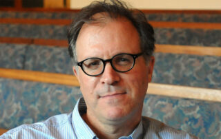 A middle-aged man with short brown hair and round black glasses is sitting indoors. He is wearing a blue-striped shirt and is seated in front of empty, patterned benches.