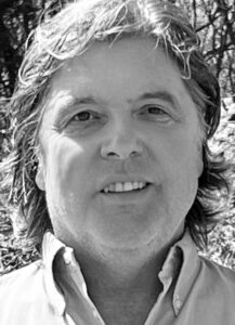 A middle-aged man with wavy hair smiles at the camera in an outdoor setting. He is wearing a button-up shirt and the photo is in black and white. Trees and foliage are visible in the background.