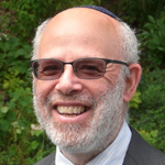 A smiling older man with a gray beard and glasses, wearing a dark suit and a kippah, standing outdoors with greenery in the background.